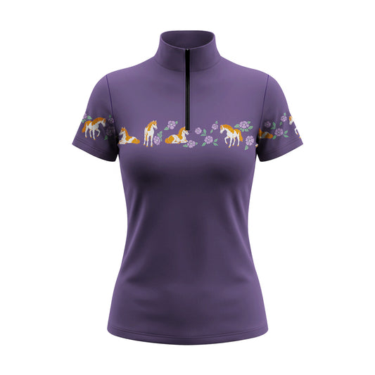 Outdoor stone‑building lifestyle shot of female equestrian wearing purple vintage pony and purple floral border‑print short‑sleeve quarter‑zip riding top and white riding trousers with brown leather belt