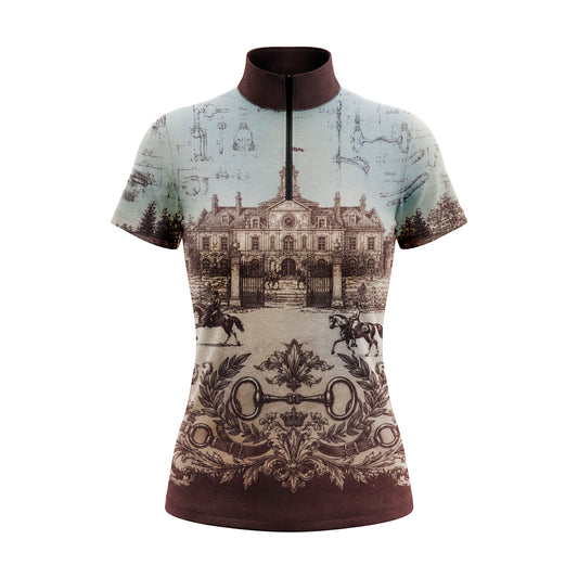White fence paddock lifestyle shot of female equestrian leaning on fence wearing vintage French chateau blueprint print short‑sleeve riding top, paired with white breeches