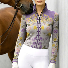 Model wearing lavender floral riding top beside chestnut horse leaning against grey wall