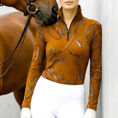 Model wearing the RIDER ATELIER Equestrian Tack Print Long-Sleeve Riding Top at the stable, with a chestnut horse, highlighting the print’s watercolor leather and metal details