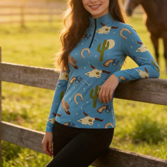 Model wearing the RIDER ATELIER Western Cowboy Print Riding Top leaning against a wooden fence at sunset