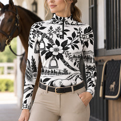 Rider in the printed top standing in a stable aisle with a horse and tack visible nearby