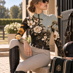 Seated rider wearing the floral riding top next to a riding helmet and saddle pad by a stable door