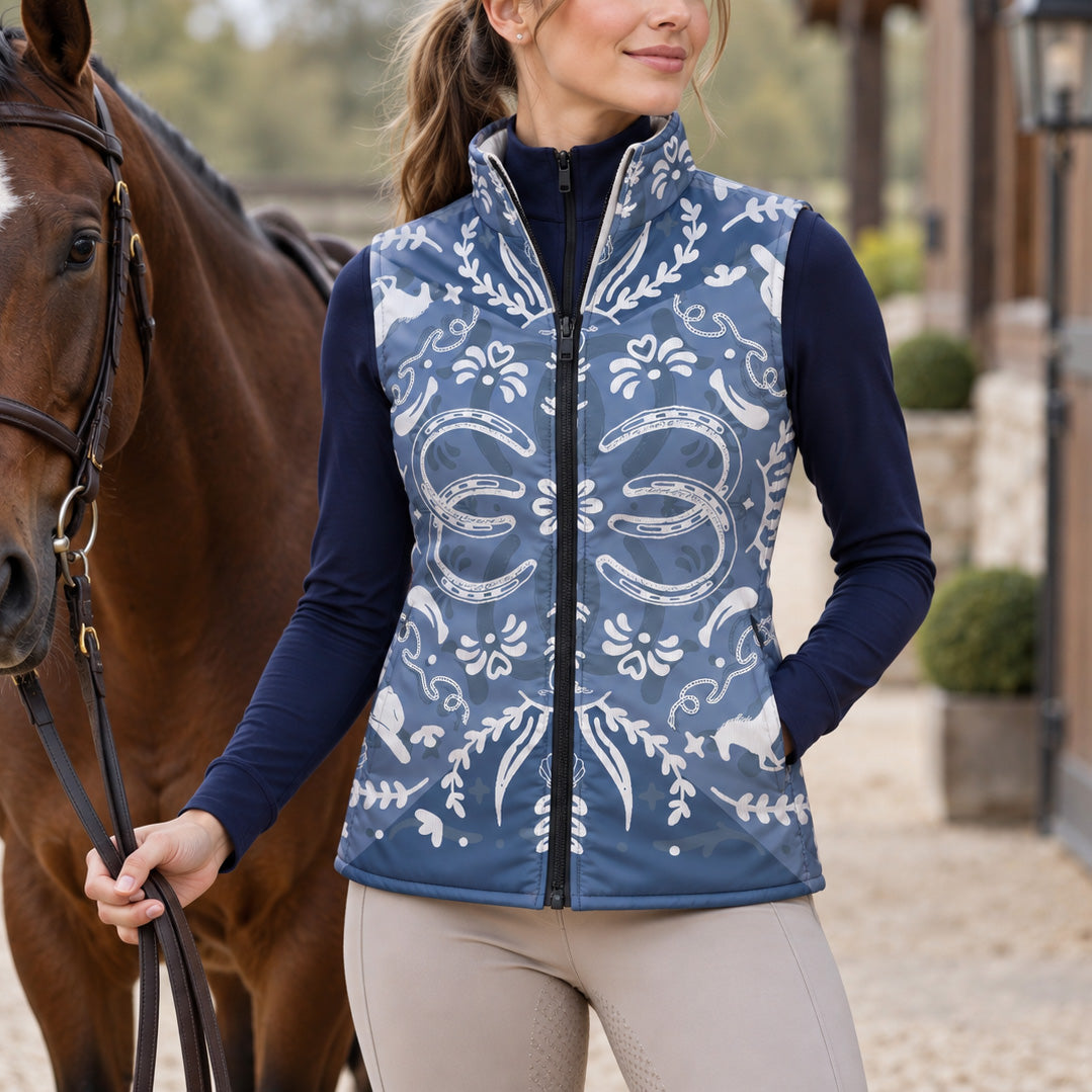 Woman wearing the blue horse and horseshoe print side of a reversible quilted vest over a navy long sleeve top beside a brown horse