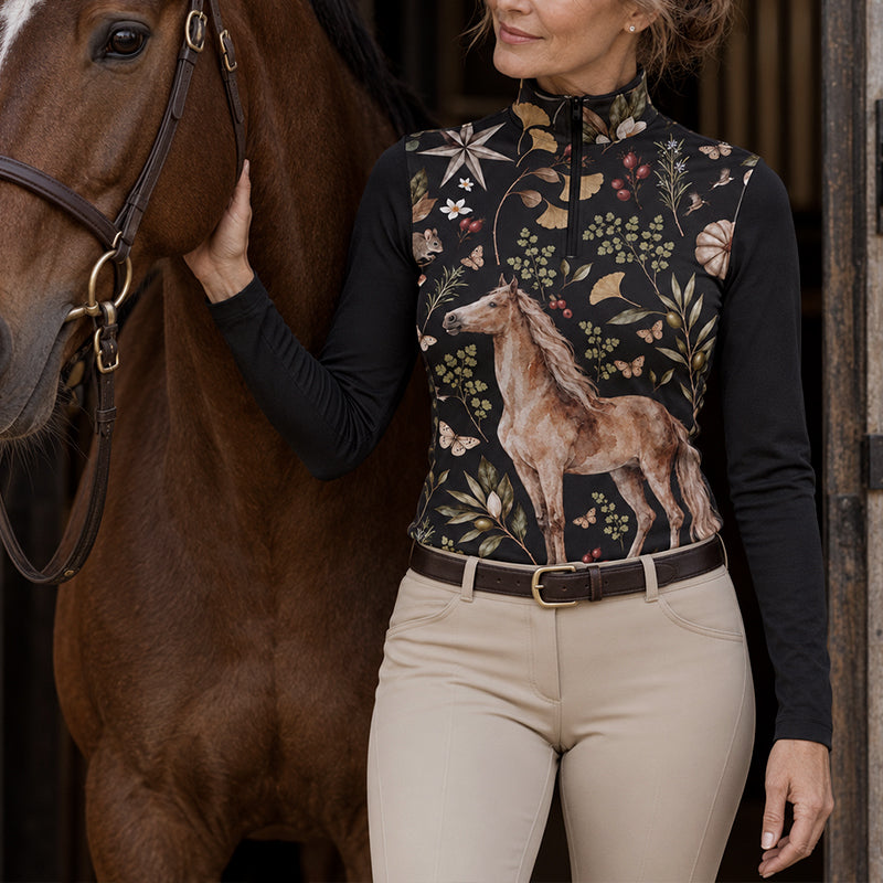 Woman wearing the black quarter zip riding top with a horse and botanical print while holding a riding crop