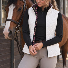 Woman wearing a white diamond quilted reversible vest over a black long sleeve top with tan breeches and tall brown boots beside a horse
