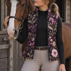 Dark horse and floral print side of a reversible equestrian vest worn over a black long sleeve top near horses and stable structures