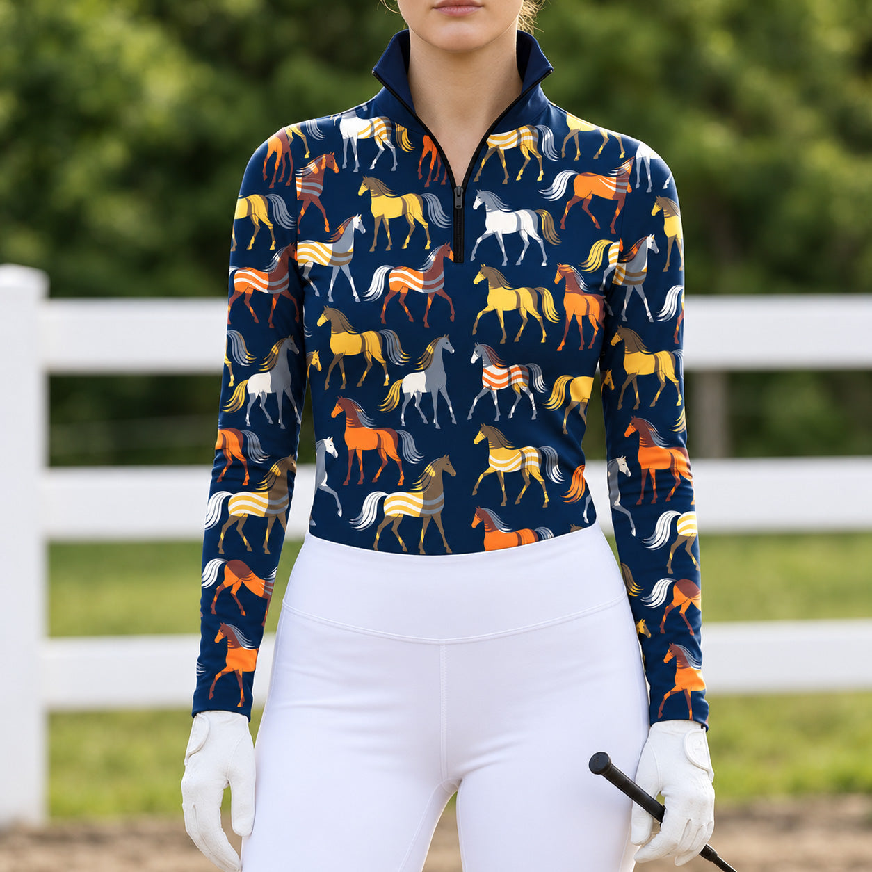 Woman wearing a navy blue horse print long sleeve quarter zip riding top with white breeches, white gloves, and a black crop in an outdoor arena with a white fence