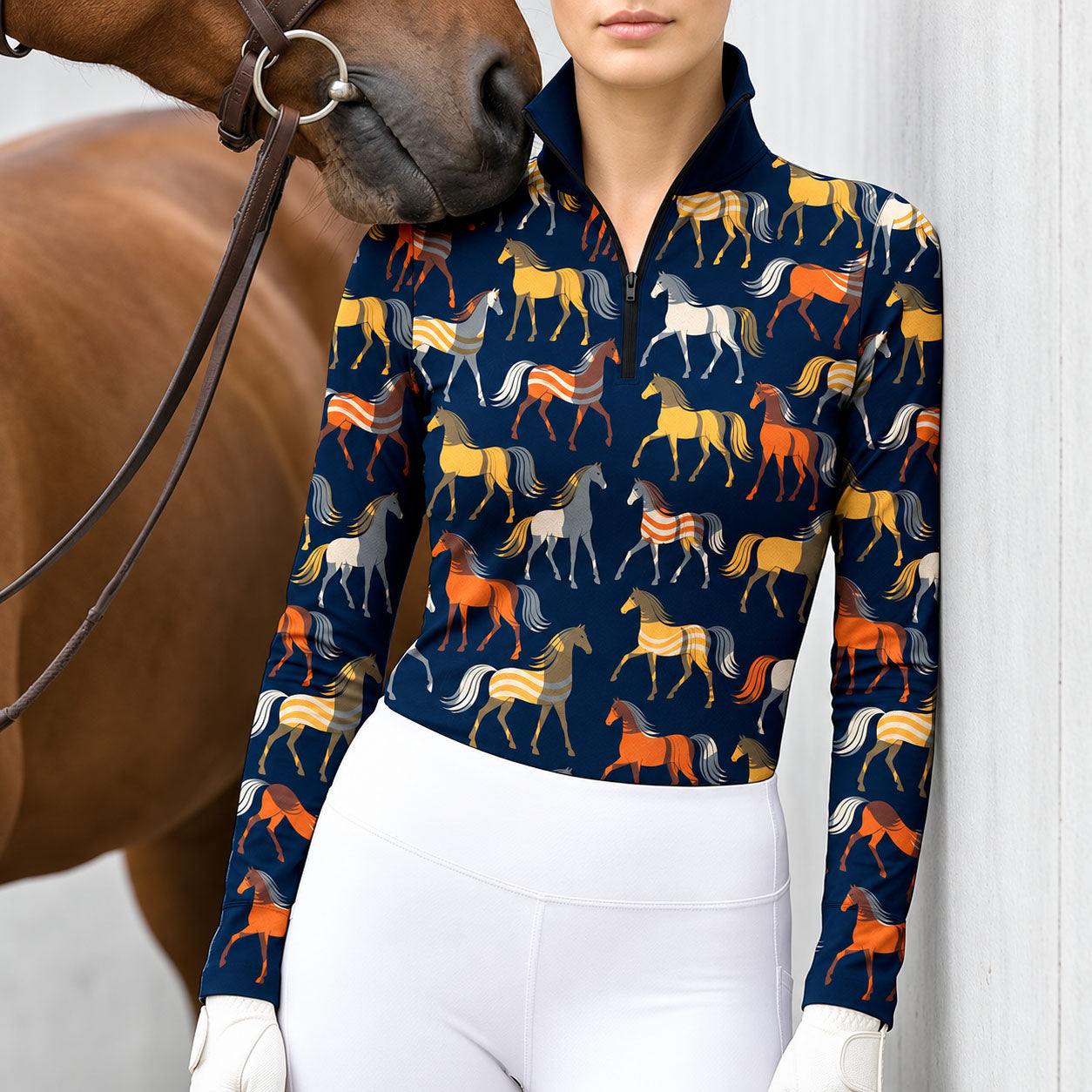 Woman wearing a navy blue horse print long sleeve quarter zip riding top with white breeches and white gloves, next to a brown horse with a leather bridle against a white wall