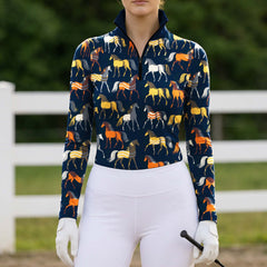 Navy blue horse print long sleeve quarter zip riding top styled with white breeches, white gloves, and a black crop in an outdoor arena with a white fence and green foliage