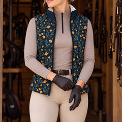 Model in a tack room wearing the navy floral print vest open over a taupe quarter zip top, beige breeches, brown leather belt, and dark brown leather riding gloves