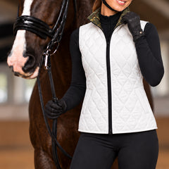 Woman wearing the white diamond quilted side of the reversible vest over a black long sleeve base layer with black breeches and gloves in an indoor arena