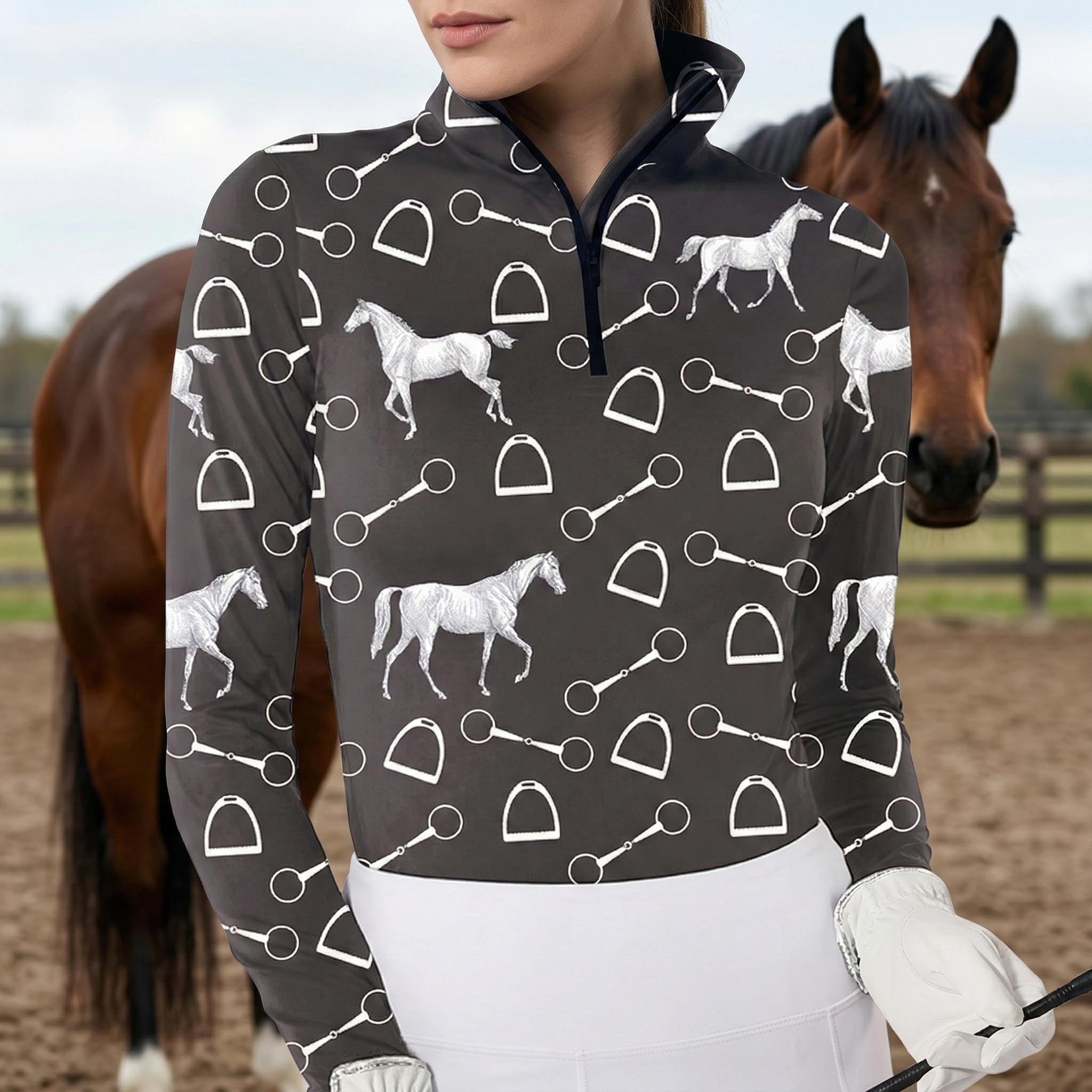 Woman wearing black equestrian silver horse and tack print long sleeve zip top standing beside a chestnut horse at the stable