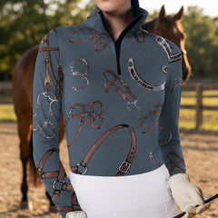 Woman wearing slate blue equestrian bridle and horseshoe print long sleeve zip top standing beside a horse at the stable