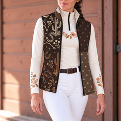 Woman wearing a brown reversible quilted vest with western artwork and light diamond shapes over a white quarter zip top and white riding pants
