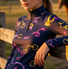 Outdoor fence‑side lifestyle shot of female equestrian wearing navy‑base long‑sleeve riding top with golden bit buckle and pink purple ribbon all‑over print