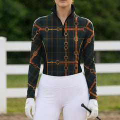 Outdoor white fence full‑front lifestyle shot of female equestrian wearing dark green tartan long‑sleeve riding top with vintage leather‑strap bridle hardware print