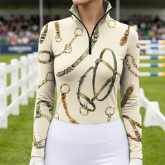 Outdoor competition show‑ground lifestyle shot of female equestrian wearing cream off‑white long‑sleeve riding top with bridle‑and‑bit hardware print