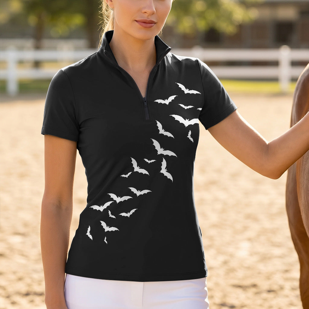 Outdoor equestrian arena lifestyle shot of female rider wearing black Halloween short‑sleeve riding top with white bat print, standing beside horse in white breeches