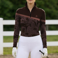 Outdoor white fence full‑front lifestyle shot of female equestrian wearing two‑tone brown long‑sleeve quarter‑zip riding top with equestrian‑tack silhouette collage print