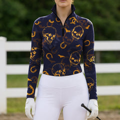 Outdoor paddock show‑ground lifestyle shot of female equestrian wearing navy long‑sleeve riding top with golden skull bat horseshoe all‑over print