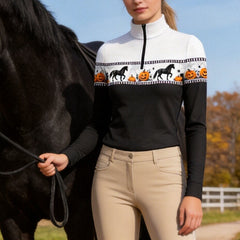 Autumn outdoor pasture lifestyle shot of female equestrian holding lead rope beside black horse wearing black‑body Halloween horse jack‑o‑lantern print long‑sleeve quarter‑zip riding top and beige breeches