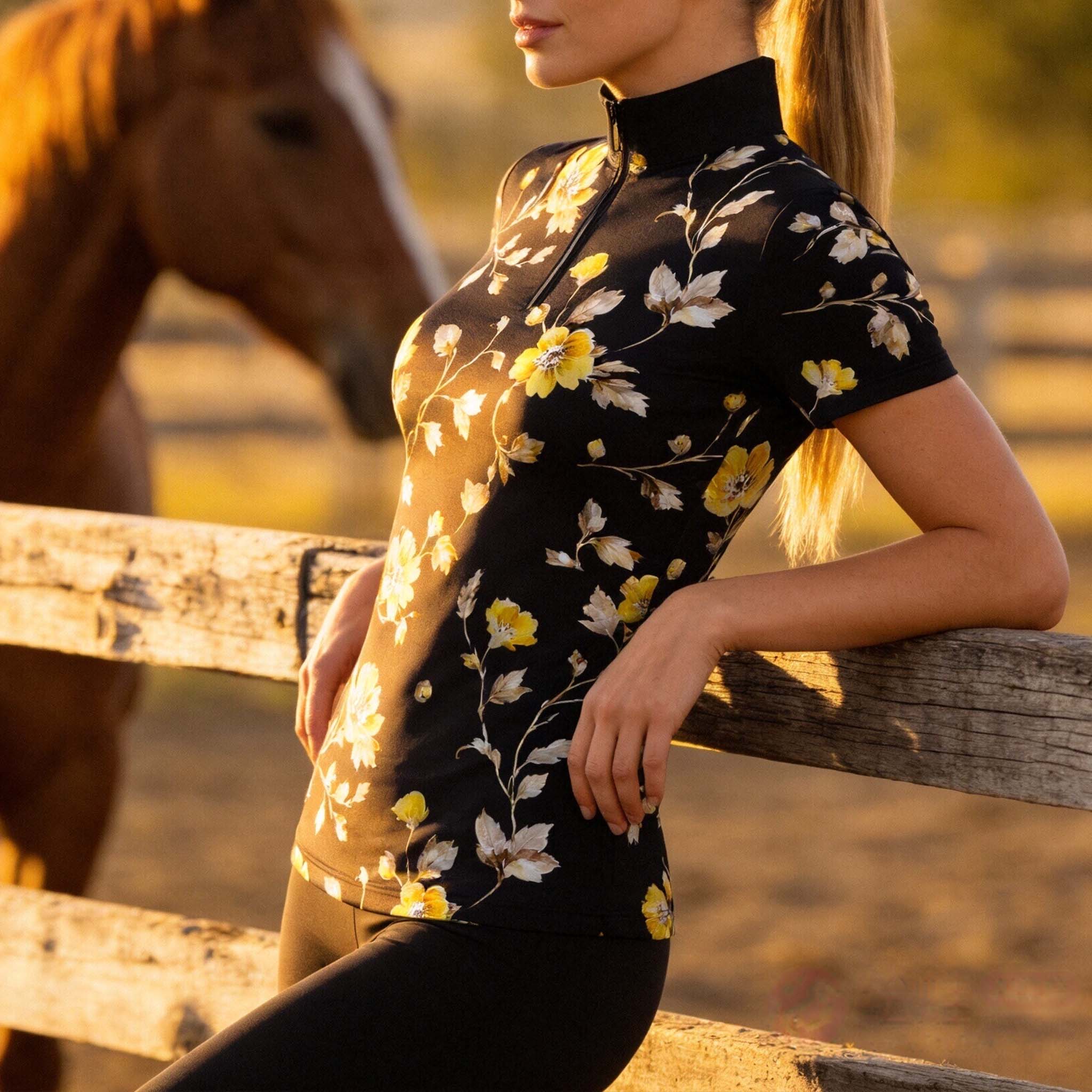 Sunset ranch fence lifestyle shot of female equestrian wearing black watercolor golden‑yellow floral print short‑sleeve riding top with horse in background