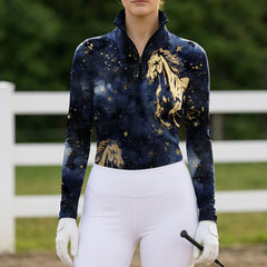 Outdoor white fence arena lifestyle shot of female equestrian wearing navy galaxy themed long‑sleeve riding top with golden galloping horse all‑over print