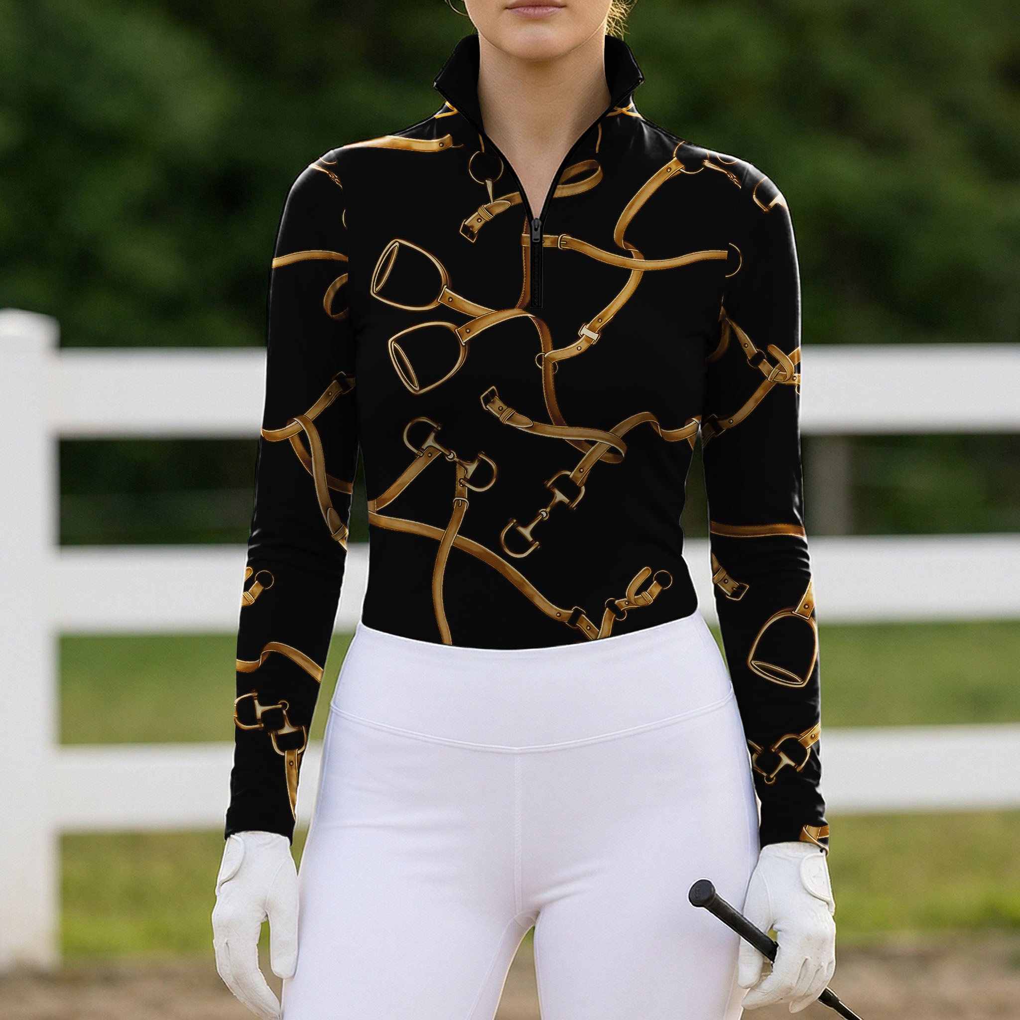 White‑fence arena lifestyle shot of female rider in black golden equestrian‑tack‑print long‑sleeve riding top with riding gloves