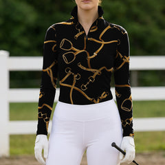 White‑fence arena lifestyle shot of female rider in black golden equestrian‑tack‑print long‑sleeve riding top with riding gloves