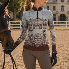 Outdoor arena lifestyle shot of female equestrian wearing burgundy‑trim vintage French chateau blueprint‑art print long‑sleeve riding top paired with taupe riding breeches, standing beside brown horse