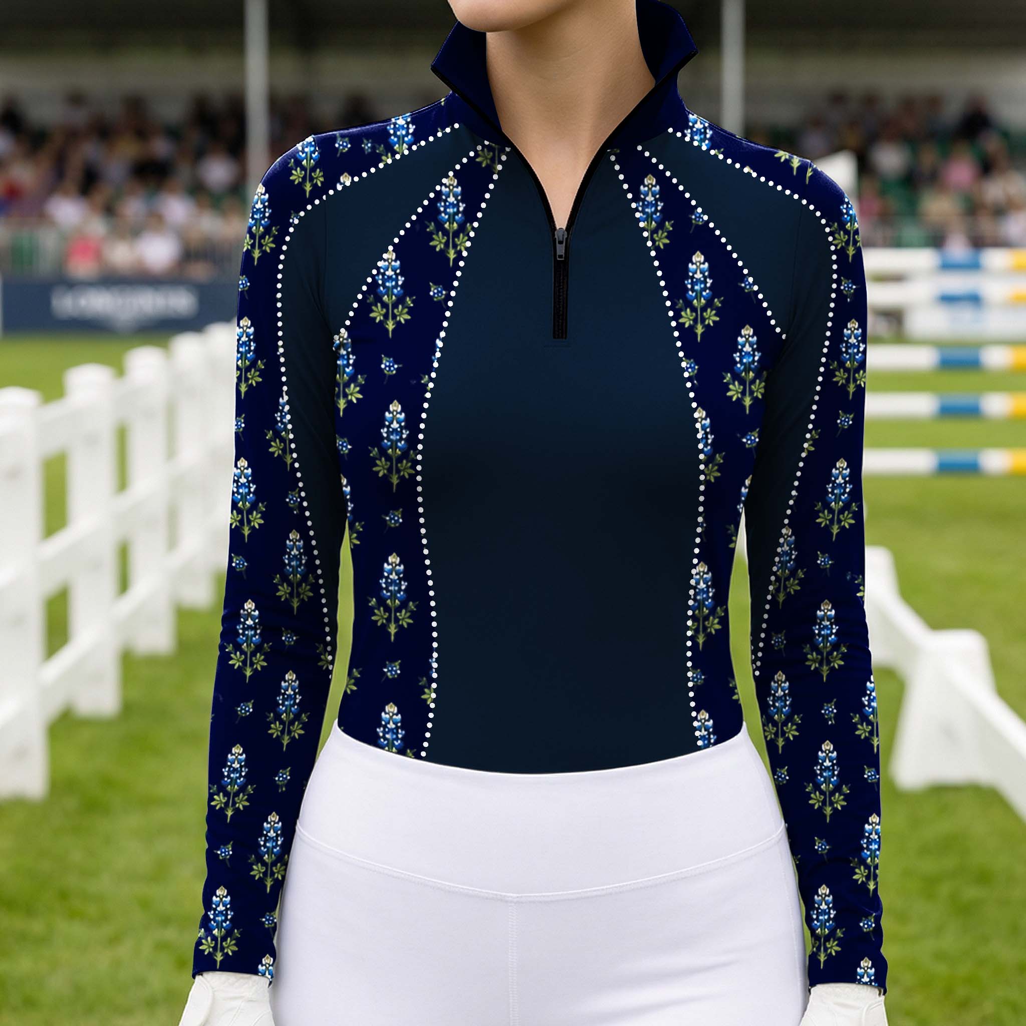 Outdoor equestrian show arena shot of female rider wearing navy bluebonnet print rhinestone‑trim long‑sleeve riding top, paired with white breeches and white riding gloves