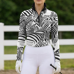 Outdoor riding arena lifestyle shot of female rider wearing zebra collage print long‑sleeve riding top, white breeches and white riding gloves holding riding crop