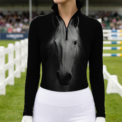 Outdoor show‑arena lifestyle shot of female equestrian wearing black long‑sleeve quarter‑zip riding top with large realistic monochrome white horse‑head front print