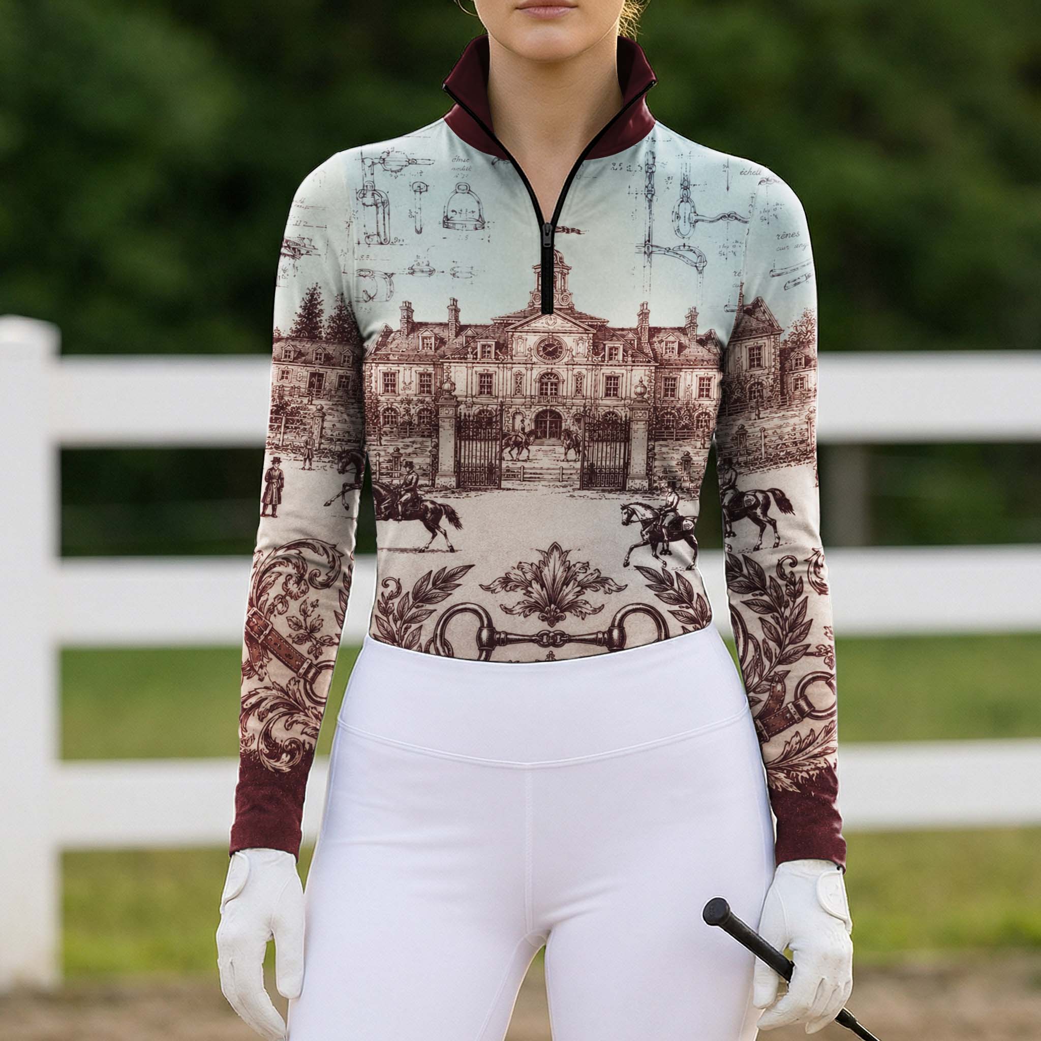 White‑fence paddock lifestyle shot of female equestrian wearing burgundy‑trim vintage French chateau blueprint‑art long‑sleeve riding top paired with clean white riding breeches and white riding gloves