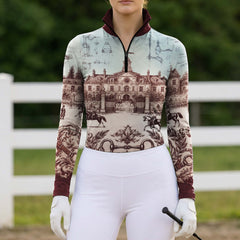 White‑fence paddock lifestyle shot of female equestrian wearing burgundy‑trim vintage French chateau blueprint‑art long‑sleeve riding top paired with clean white riding breeches and white riding gloves