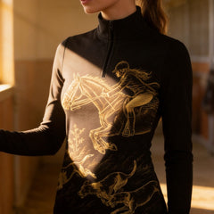 Warm sunlit stable lifestyle shot of equestrian wearing black long‑sleeve riding top with gold jumping rider and hounds graphic