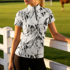 Sandy riding arena front‑angle lifestyle shot of female equestrian standing beside horse wearing white short‑sleeve riding top with black sketch galloping horse all‑over print
