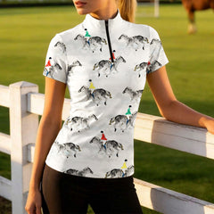 Golden‑hour green pasture lifestyle shot of female equestrian leaning on white wooden fence wearing white short‑sleeve riding top with watercolor jumping rider all‑over print