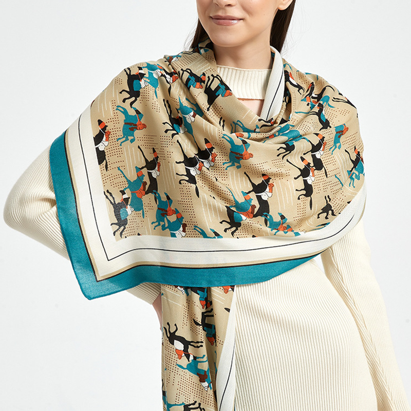 A woman wearing a cream ribbed sweater and a large beige scarf with a colorful print of stylized figures and a teal border, photographed against a light gray studio background.
