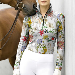Lifestyle shot with brown horse nuzzling rider’s shoulder, female equestrian leans against light wall in cream watercolor wildflower unicorn‑pony print riding top and white riding gloves