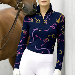 Stable‑wall lifestyle shot of female equestrian beside horse wearing navy‑base long‑sleeve riding top with golden bit buckle and pink purple ribbon all‑over print