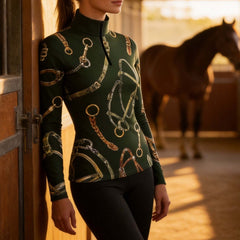 Warm‑toned indoor stable lifestyle shot of female equestrian leaning against wooden stall door wearing forest‑green long‑sleeve riding top with bridle‑and‑bit print