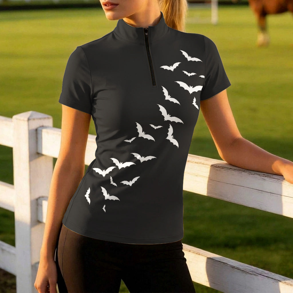 Sunny pasture fence lifestyle shot of woman rider in black Halloween bat‑print short‑sleeve riding top and dark riding pants leaning against white wooden fence