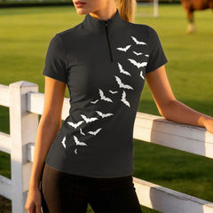 Sunny pasture fence lifestyle shot of woman rider in black Halloween bat‑print short‑sleeve riding top and dark riding pants leaning against white wooden fence