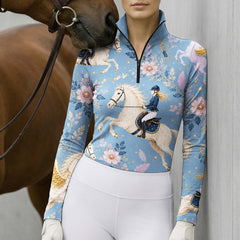 Outdoor wall lifestyle shot of female equestrian with brown horse nuzzling shoulder wearing dusty‑blue fairytale pegasus print long‑sleeve riding top with white gloves and white breeches