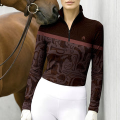 Outdoor wall lifestyle shot of female equestrian standing beside brown horse wearing two‑tone brown long‑sleeve quarter‑zip riding top with equestrian‑tack collage artwork