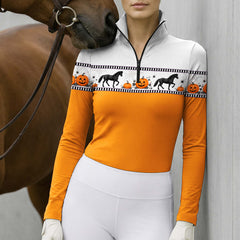Outdoor paddock lifestyle shot of female rider beside brown horse wearing white gloves in orange‑body Halloween horse‑pumpkin border‑print long‑sleeve riding top paired with white breeches