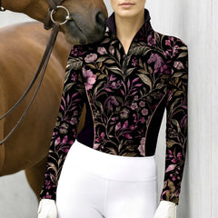 Lifestyle shot of female equestrian in black floral horse‑silhouette long‑sleeve riding top standing beside brown horse against light‑colored wall, wearing white breeches and gloves