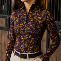 Stall close‑up lifestyle shot of female equestrian wearing deep‑purple golden abstract floral‑geometric doodle print long‑sleeve riding top and light riding breeches with leather belt